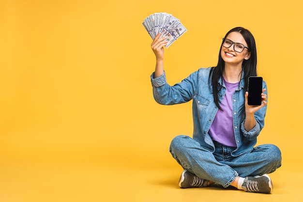 Feliz vencedor Retrato de uma mulher alegre sorridente mostrando smartphone em branco isolado sobre fundo amarelo Sentado no chão em pose de lótus segurando notas de dinheiro