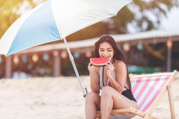 Feliz verano y vacaciones, vacaciones, fin de semana largo en la playa. Mujer sexy relajante vistiendo bikini sentada en una silla de playa y fruta tropical de sandía, sonríe y relájate