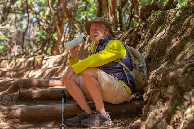 Feliz viajante idoso com boné e mochila desfrutando de caminhada na natureza sentado na floresta bebendo de sua garrafa de água