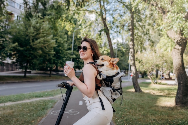 Feliz viajante sorridente está de pé com eletro scooter e bebendo café no parque da cidade com cachorro Welsh Corgi Pembroke em uma mochila especial