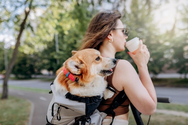 Feliz viajante sorridente está de pé com eletro scooter e bebendo café no parque da cidade com cachorro Welsh Corgi Pembroke em uma mochila especial