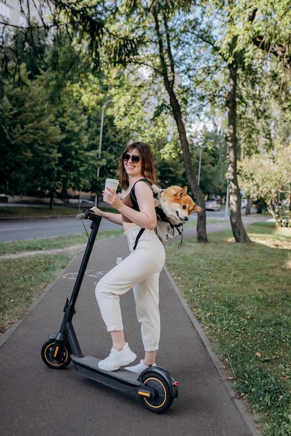 Feliz viajante sorridente está de pé com eletro scooter e bebendo café no parque da cidade com cachorro Welsh Corgi Pembroke em uma mochila especial