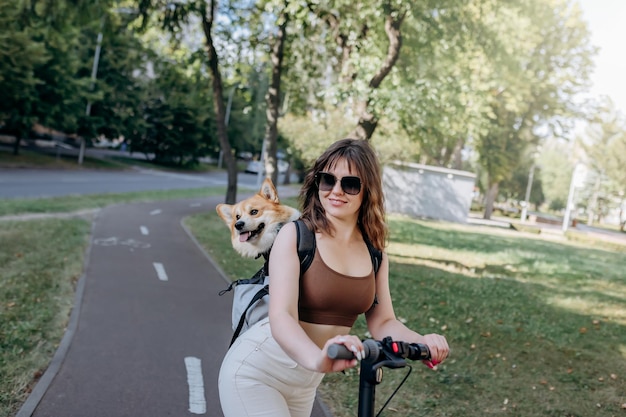 Feliz viajante sorridente está montando sua eletro scooter no parque da cidade com cachorro welsh corgi pembroke em uma mochila especial