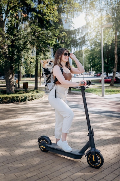 Feliz viajante sorridente está montando sua eletro scooter no parque da cidade com cachorro Welsh Corgi Pembroke em uma mochila especial