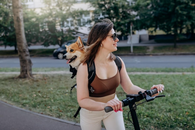 Feliz viajante sorridente está montando sua eletro scooter no parque da cidade com cachorro Welsh Corgi Pembroke em uma mochila especial