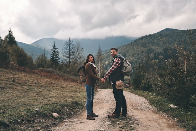 Feliz de viajar juntos. Longitud total de pareja joven tomados de la mano y sonriendo mientras está de pie en el sendero en las montañas