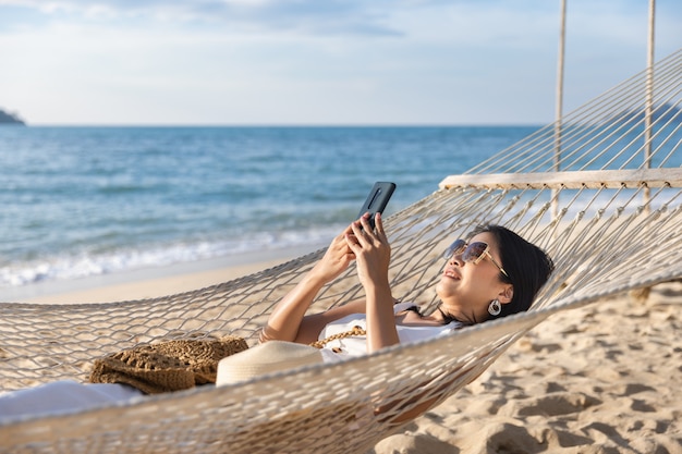 Feliz viajero mujer asiática con teléfono móvil y relajarse en una hamaca en la playa en Koh Chang, Trad, Tailandia