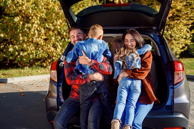 Felizes alegres jovens pais sentados no porta-malas do carro e abraçam seus filhos felizes correndo para eles.