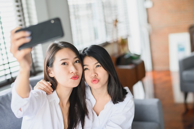 Felizes amigas asiáticas usando smartphone selfie na sala de estar