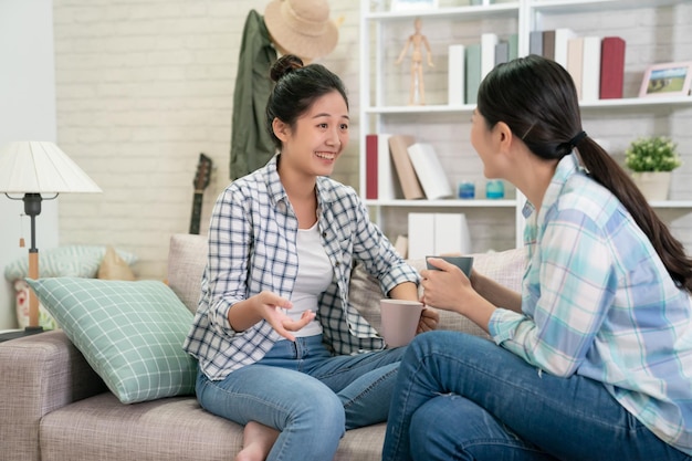 Felizes amigas japonesas asiáticas em casa sentadas no sofá. duas jovens com café conversando no sofá fofocando e compartilhando segredos discutindo a vida e as relações. conceito de confiança de amizade