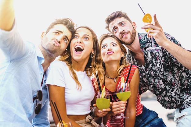 Felizes amigos bêbados tirando uma selfie fazendo caretas segurando coquetéis alcoólicos sentados em um banco ao ar livre no amor de amizade de verão no happy hour e conceito de estilo de vida de álcool