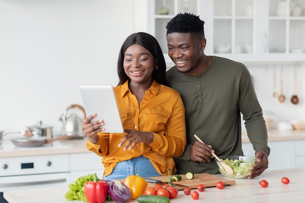 Felizes cônjuges afro-americanos preparando comida na cozinha e usando tablet digital