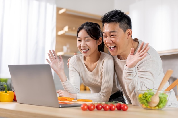 Felizes cônjuges asiáticos fazendo videochamada enquanto cozinham na cozinha