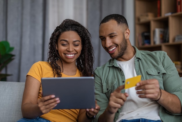 Felizes cônjuges negros comprando on-line usando tablet e cartão de crédito pagando por pedidos sentados no sofá em