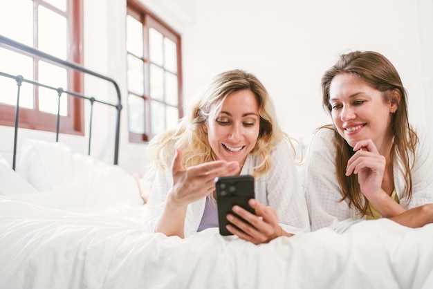 Felizes duas amigas, jovem caucasiana em roupões de banho brancos, usando um smartphone junto com diversão e sorrisos enquanto estava deitada na cama em casa, casais lgbtq, spa de lazer e atividades de tratamento