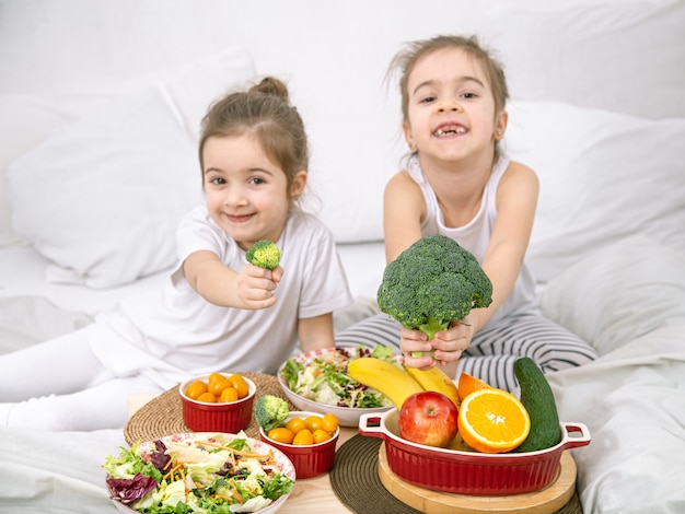 Felizes duas garotas bonitas comendo frutas e vegetais no quarto na cama. Alimentação saudável para crianças e adolescentes.