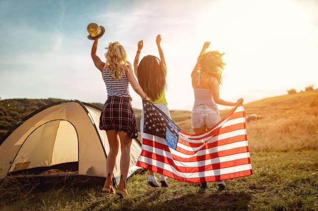 Felizes jovens amigas desfruta de um dia ensolarado na natureza. Eles estão dançando e segurando uma bandeira americana na frente de uma barraca de acampamento. Visão traseira.