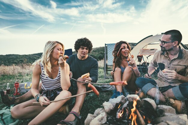 Felizes jovens amigos desfrutam de um bom dia na natureza. Eles estão grelhando salsichas, rindo e conversando felizes por estarem juntos.