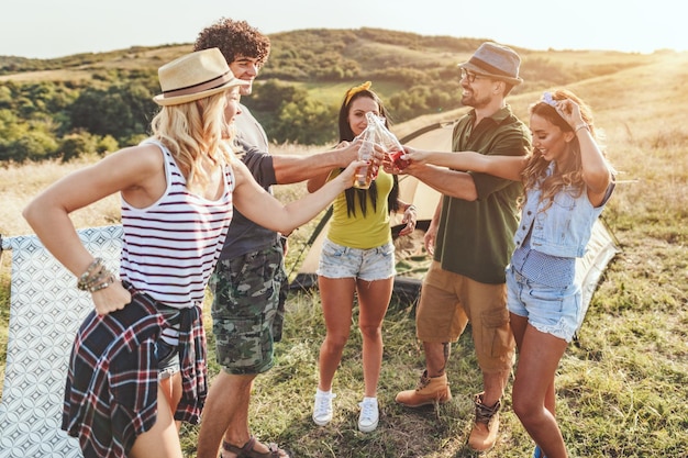 Felizes jovens amigos desfrutam de um dia ensolarado na natureza. Eles estão brindando com garrafas de cerveja, rindo e conversando felizes por estarem juntos.