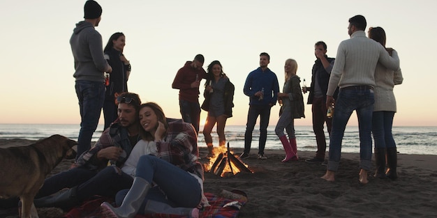 Felizes jovens amigos despreocupados se divertindo e bebendo cerveja por bonefire na praia enquanto o sol começa a se pôr