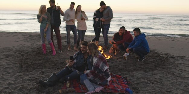 Felizes jovens amigos despreocupados se divertindo e bebendo cerveja por Bonefire na praia enquanto o sol começa a se pôr