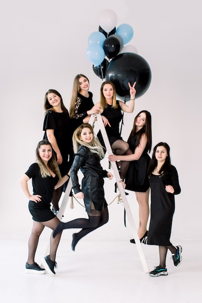 Felizes jovens sete garotas de vestido preto se divertindo comemorando aniversário na sala de luz. os amigos se divertindo posando na escada, decorada com balões de ar coloridos.