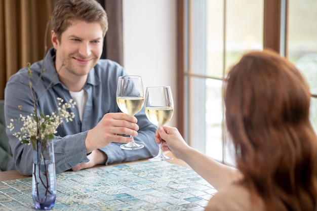 Felizes juntos. jovem adulto sorridente com uma camisa cinza estendendo a mão com um copo de vinho para uma mulher sentada de costas para a câmera