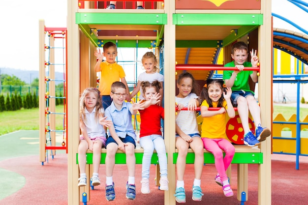 Felizes meninos e meninas de diferentes idades sentados juntos em construções de madeira e metal no playground ao ar livre, acenando, olhando para a câmera