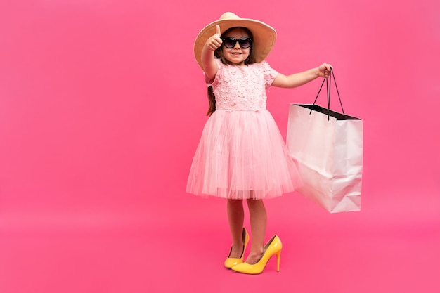 Felizes momentos adoráveis de fazer compras com a menina bonitinha no vestido de pé nos sapatos grandes da mãe com pacotes brancos nas mãos isoladas no fundo rosa.
