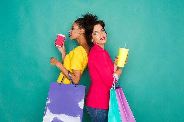Felizes namoradas multiétnicas com sacolas de compras e bebidas para levar. Dois shopaholics animados em um fundo turquesa de sudio com espaço de cópia