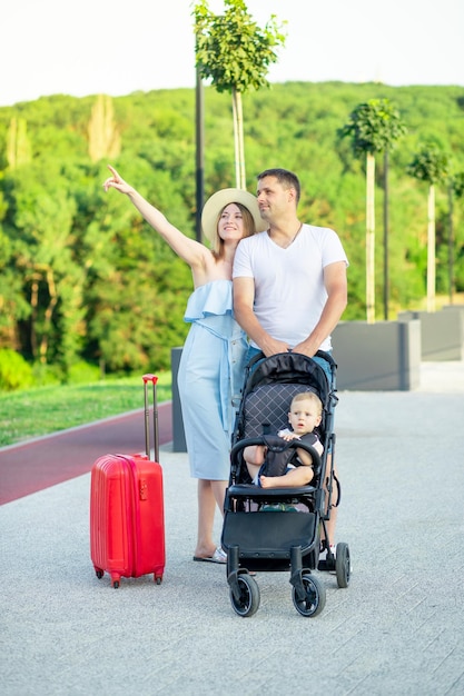 Foto felizes pais jovens mãe e pai com uma mala vermelha e um bebê em um carrinho estão indo em uma viagem de férias no verão apontando para a distância