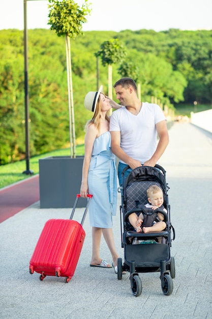 Felizes pais jovens mãe e pai com uma mala vermelha e um bebê em um carrinho estão indo em uma viagem de férias no verão sorrindo