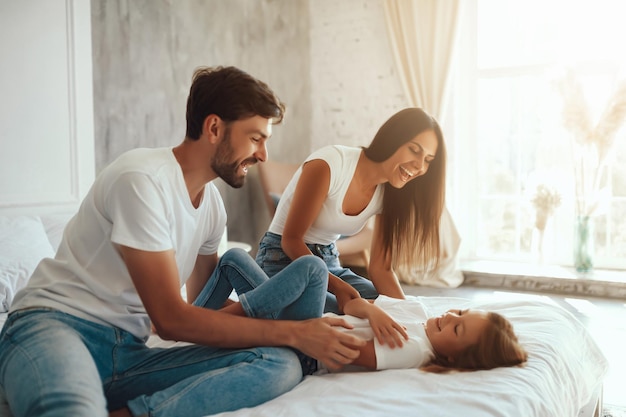 Felizes pais jovens se divertindo com sua filhinha enquanto ficam dentro de casa e deitados em uma cama