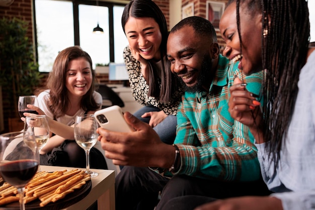 Felizes várias nacionalidades, amigos de grupo de etnias olhando, assistindo mensagens engraçadas na tela do smartphone, sorrindo, rindo, bebendo vinho, comendo palitos de pão na reunião do apartamento.