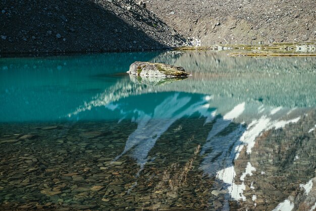 Felsen im türkisfarbenen Bergsee. Schneebedeckter Berg spiegelt sich im azurblauen klaren Wasser des Gletschersees. Schöner sonniger Hintergrund mit schneeweißer Gletscherreflexion in der grünen Wasseroberfläche des Bergsees.