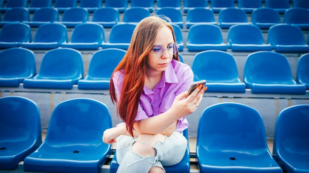 Fêmea adulta concentrada com longos cabelos castanhos na blusa e óculos, sentado no assento da tribuna azul e mensagens no smartphone