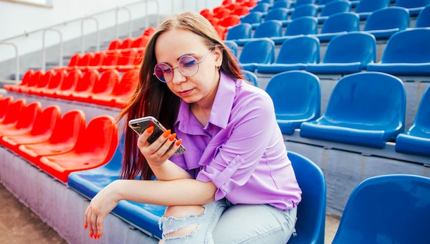 Fêmea adulta concentrada com longos cabelos castanhos na blusa e óculos, sentado no assento da tribuna azul e mensagens no smartphone