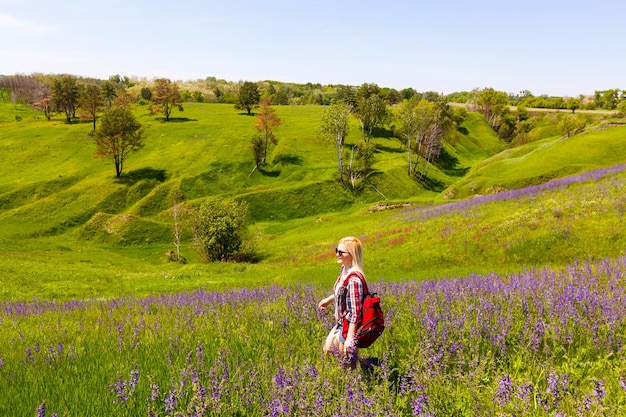 Fêmea aventureira feliz fica na encosta verde da montanha entre rododendros rosa floridos e olhando para a distância. Viagem épica nas montanhas. Ângulo amplo.