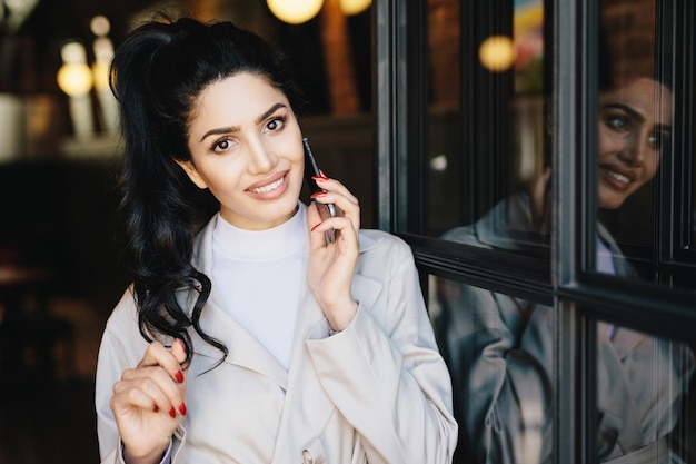 Fêmea bonita com cabelos escuros ondulados olhos encantadores lábios bem formados e dentes brancos mesmo vestindo roupas brancas com manicure vermelha se comunicando por telefone tocando seu cabelo com a mão gentil