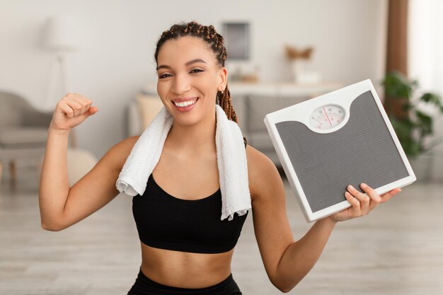 Fêmea de fitness africana feliz segurando escalas mostrando bíceps em casa