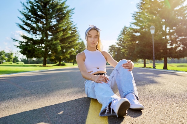 Fêmea de hipster adolescente na moda na moda sentada na estrada no parque