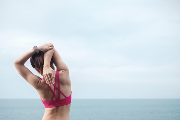 Fêmea de jovem aptidão no sportswear, alongamento do corpo contra a vista para o mar, exercício de mulher saudável de manhã. Conceitos de treino, bem-estar e equilíbrio entre vida profissional