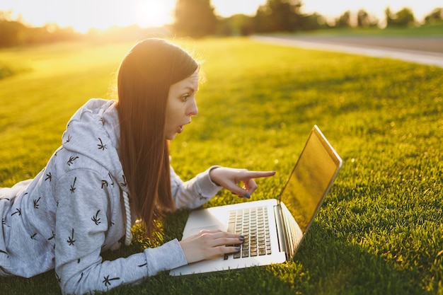 Fêmea de jovem estudante inteligente em roupas casuais. Mulher deitada no chão de grama, trabalhando no computador laptop pc no parque da cidade, no gramado do sol de grama verde ao ar livre. Escritório móvel. Conceito de negócio autônomo.