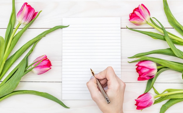 Fêmea escreve no bloco de notas em branco e tulipas na mesa de madeira branca Conceito de férias