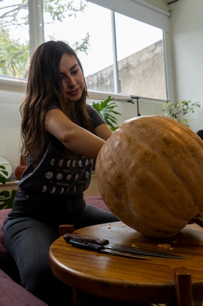 Fêmea esculpindo grande abóbora laranja para o Halloween enquanto está sentado em uma mesa de madeira em casa