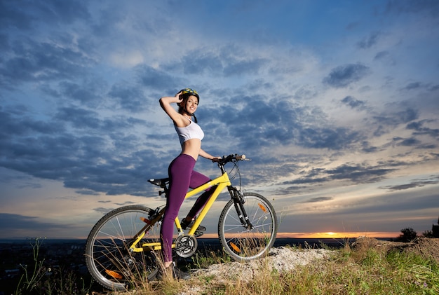 Fêmea jovem ciclista em bicicleta no topo da montanha, com bela paisagem ao pôr do sol