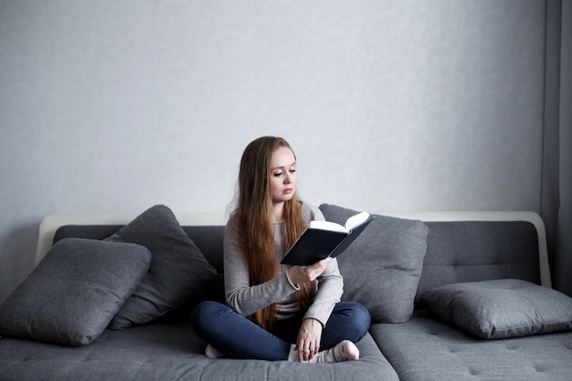 Fêmea jovem inteligente, sentado no sofá em casa lendo livro