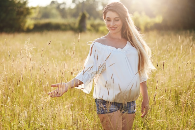 Fêmea jovem positiva antecipa bebê, passeios pelo campo de trigo, goza de dia ensolarado, toca a grama verde. Tiro ao ar livre da mulher grávida vestida com blusa branca e shorts jeans. Gravidez
