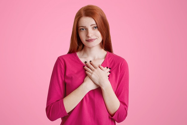 Foto fêmea sardenta amigável de gengibre mantém as mãos no coração, como expressa sua simpatia e amor, usa camisola rosa