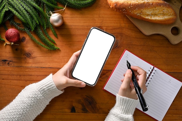 Fêmea segurando maquete de smartphone e escrevendo em seu espaço de trabalho de bloco de notas com tema de Natal